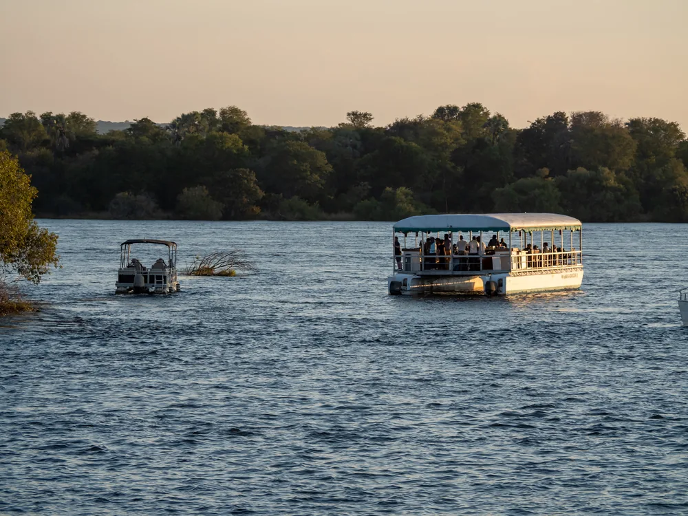 Two tourist boats on the Zambezi in golden evening light, with trees along the far bank