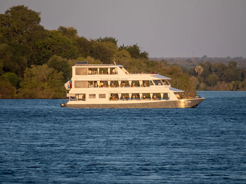 The three-deck cruise boat Zambezi Explorer, lit gold by the low sun, passing wooded banks on the Zambezi above Victoria Falls