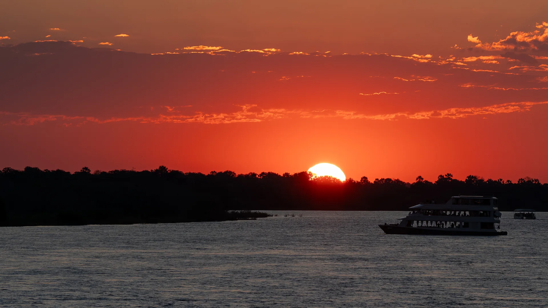 The sun setting red behind the tree line on the Zambezi River above Victoria Falls, with a two-deck cruise boat in silhouette on the water