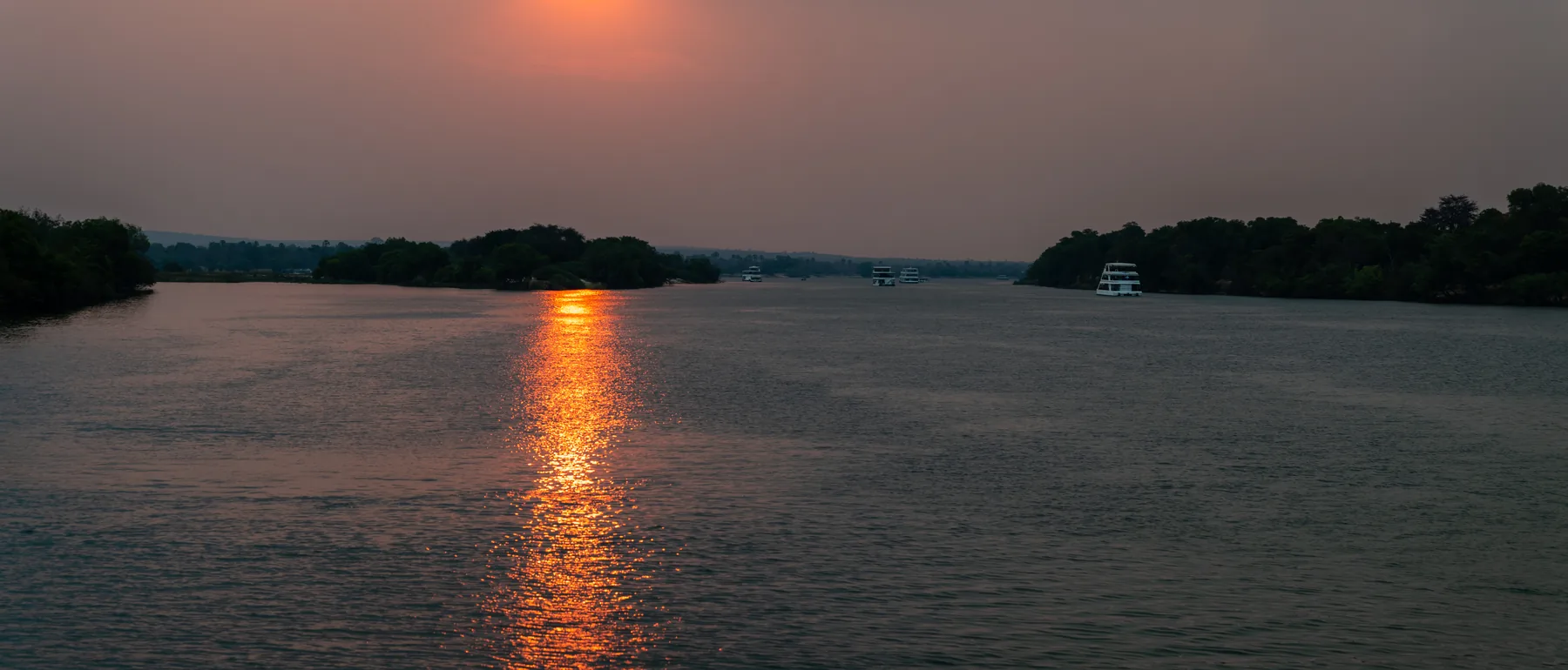 The sun low over a wide, hazy stretch of the Zambezi in Zimbabwe, its reflection running down the water between wooded islands