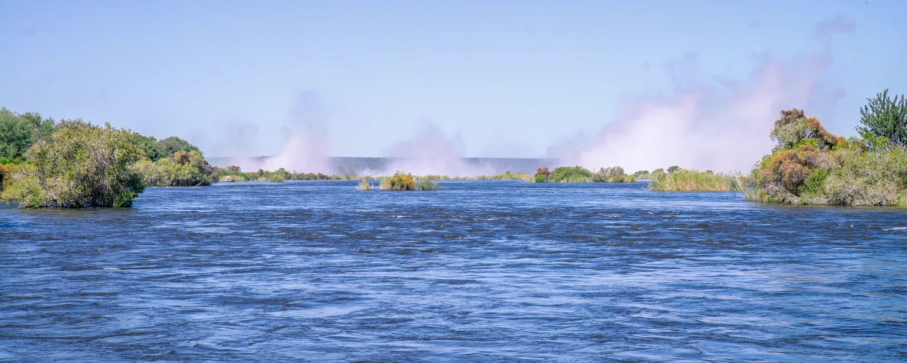 Spray from Victoria Falls rising beyond tree-covered islands on the Zambezi River upstream of the drop